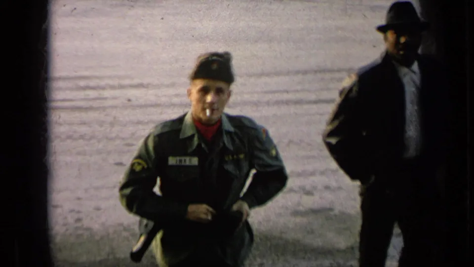 Uniformed soldier with a cigarette in his mouth looks toward the camera as a suited man in a fedora stands at the edge of frame.