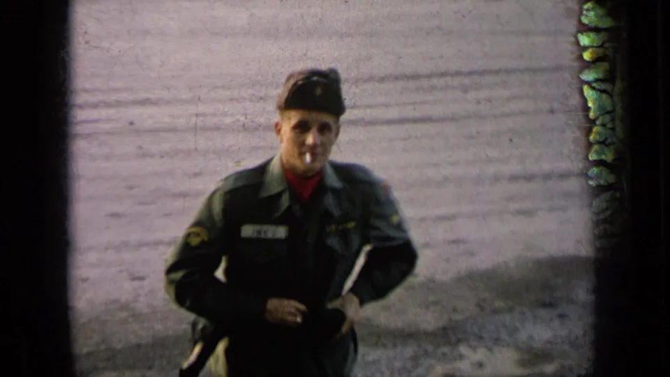 Close 8mm frame of the soldier in green uniform and cap, cigarette in mouth, standing against a plain outdoor background.