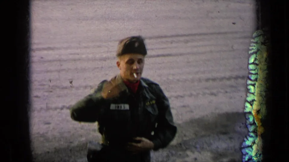 The soldier in uniform raises his hand in a brief wave while a cigarette remains in his mouth, framed by visible 8mm film edges.