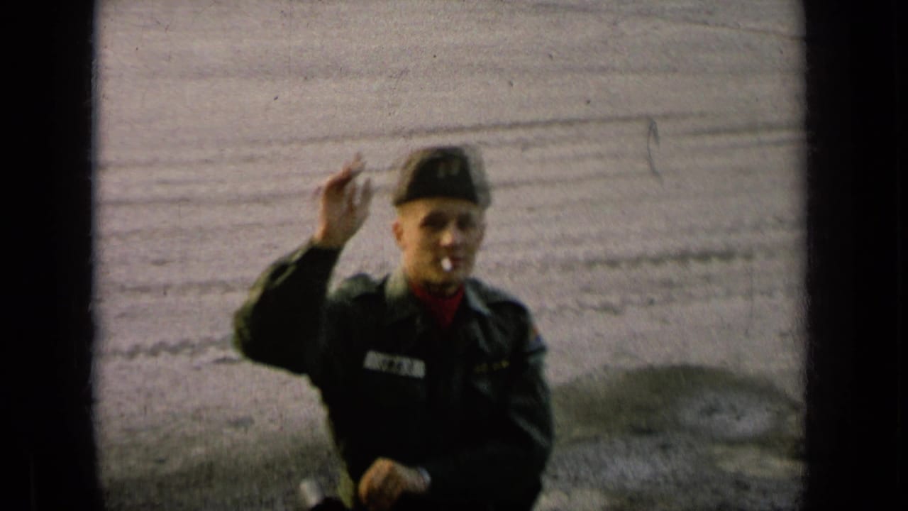Uniformed soldier in cap smokes a cigarette and raises a hand toward the camera in archival 1960s military-base footage.