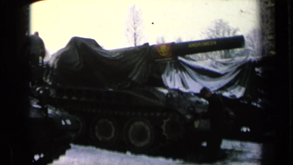 Covered tracked tank outdoors, with a painted name on the gun barrel and figures standing near the armored vehicle.
