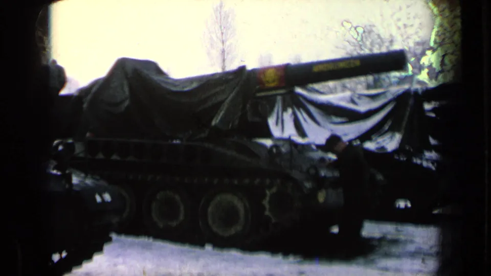 Side view of a covered tracked tank labeled on the barrel, with a person standing near the vehicle in a snowy outdoor area.