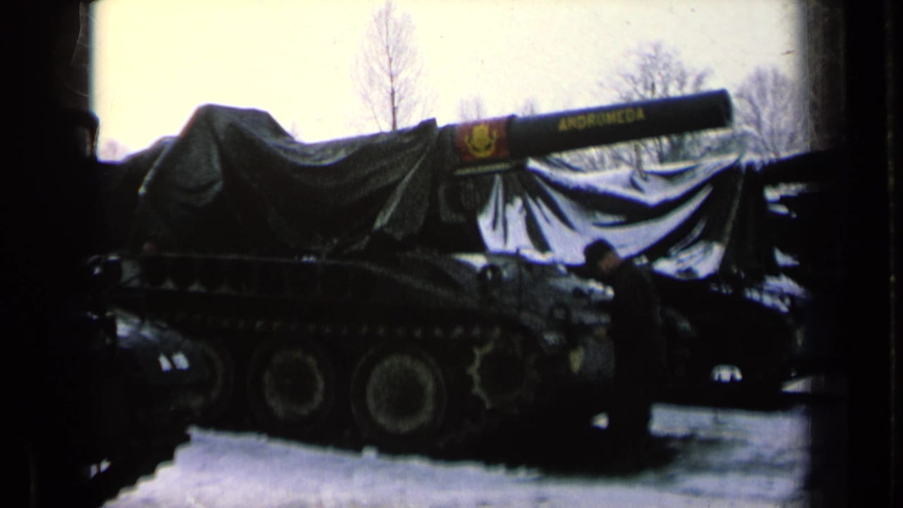 Tarp-covered tank with long barrel parked in snow beside a dark vehicle and a uniformed person.