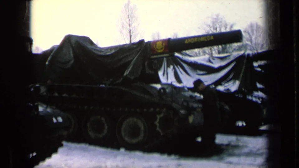 1963 8mm frame showing a covered tank on a snowy German military base, with a uniformed person near the vehicle and bare trees behind.