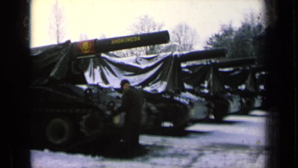 A row of covered tanks appears on a snowy military base, with one uniformed person beside the nearest tracked vehicle.