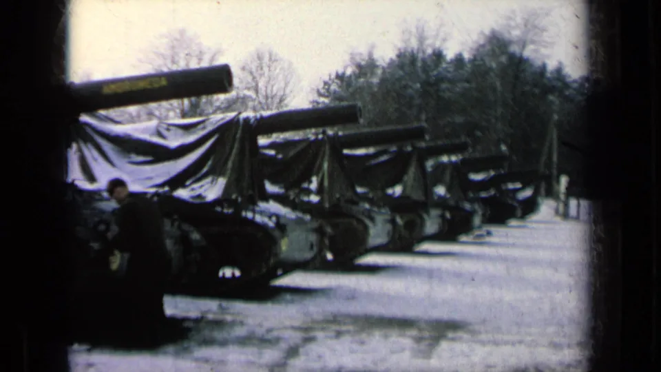 Several tarp-covered tanks are lined up outdoors in snow, with a dark-clad figure at the edge of the frame and trees in the background.