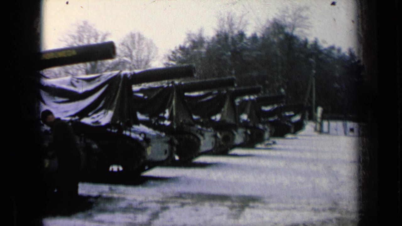 Row of tarped armored vehicles in snow with a uniformed man at left.