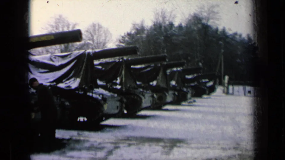 1963 8mm frame showing covered armored vehicles lined up outdoors, with a uniformed man near the left edge and bare trees behind.