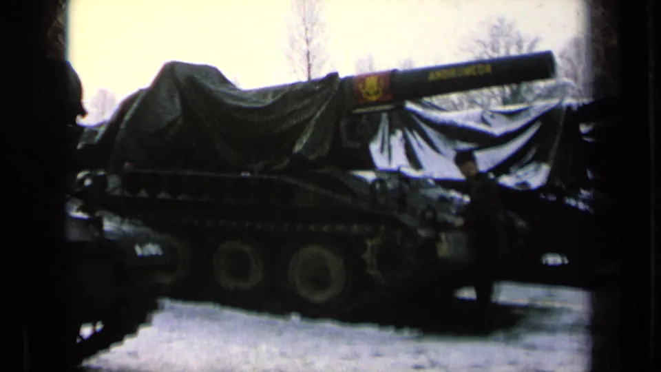 Close side view of a covered armored vehicle, with wheels visible below the tarp and a uniformed figure partly obscured near the right side.