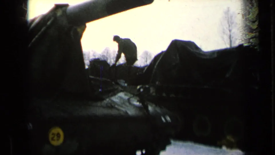 A dark 8mm frame shows a man silhouetted on or beside a tarped armored vehicle, with a large gun barrel crossing the upper frame.