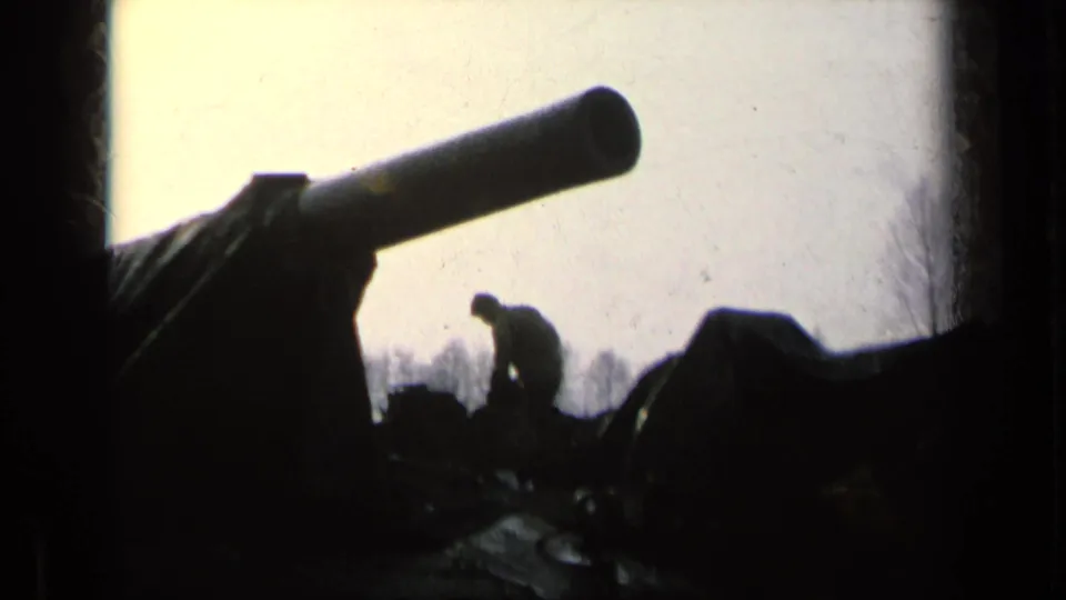 A soldier appears in silhouette beside a large tank barrel and tarp-covered armor in archival 8mm military-base footage.