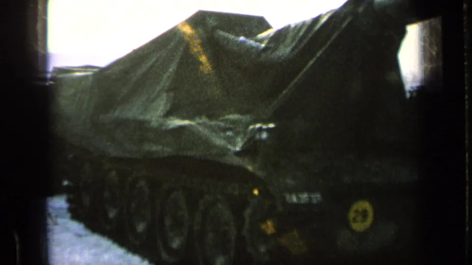 A tarp-covered tank or armored vehicle fills the frame, with wheels, small markings, and dark 8mm exposure visible.