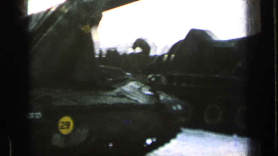 A silhouetted soldier bends near a row of tarp-covered armored vehicles, framed by dark exposure and a bright sky.