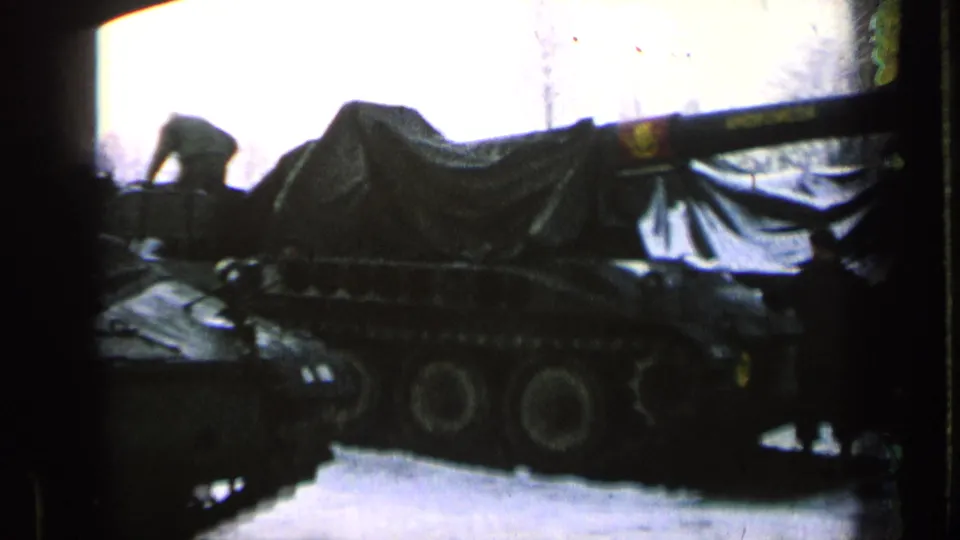 1963 8mm frame showing covered military tanks parked in snow, with a dark-clad person near the vehicles.