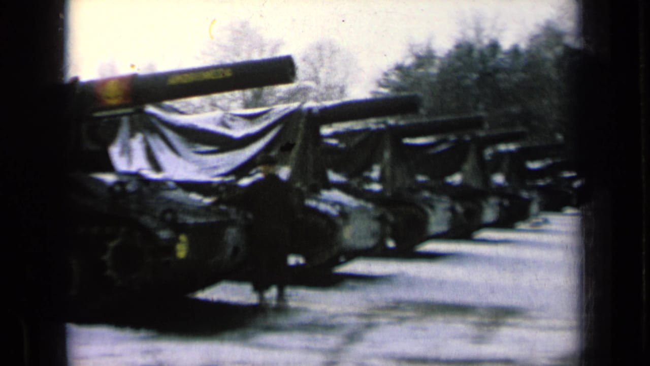 Line of covered tanks parked beside a snowy road with a person in dark clothing.