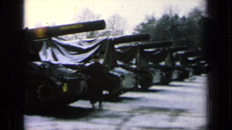 Covered military tanks line a snowy outdoor area, with one dark-clad person visible beside the vehicles.