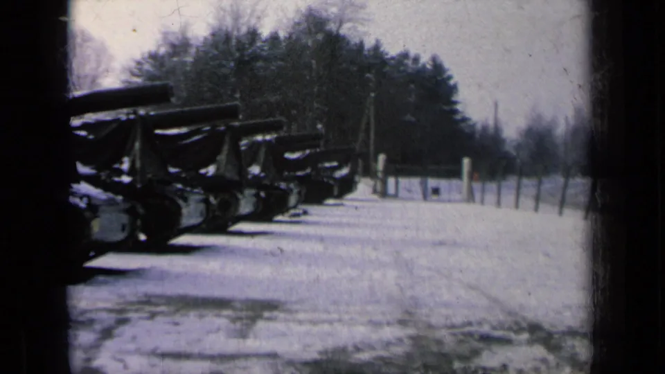 Snowy 8mm view of several covered military tanks parked in a line, with bare trees beyond the lot.