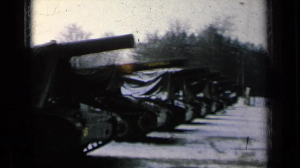 Wide 1963 8mm view of covered military tanks lined up outdoors with bare trees behind them.