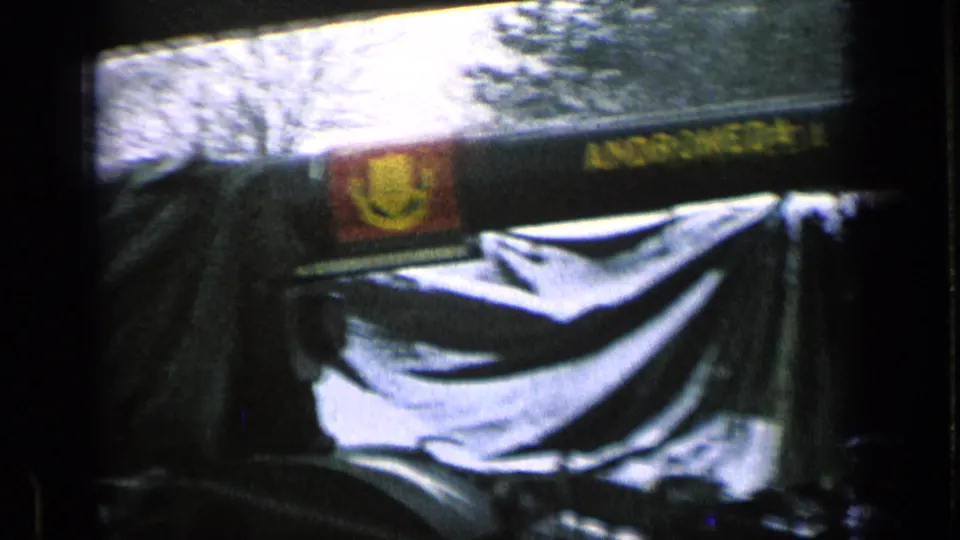 1963 8mm frame showing a row of covered armored vehicles with long gun barrels, canvas tarps, and bare trees in the background.