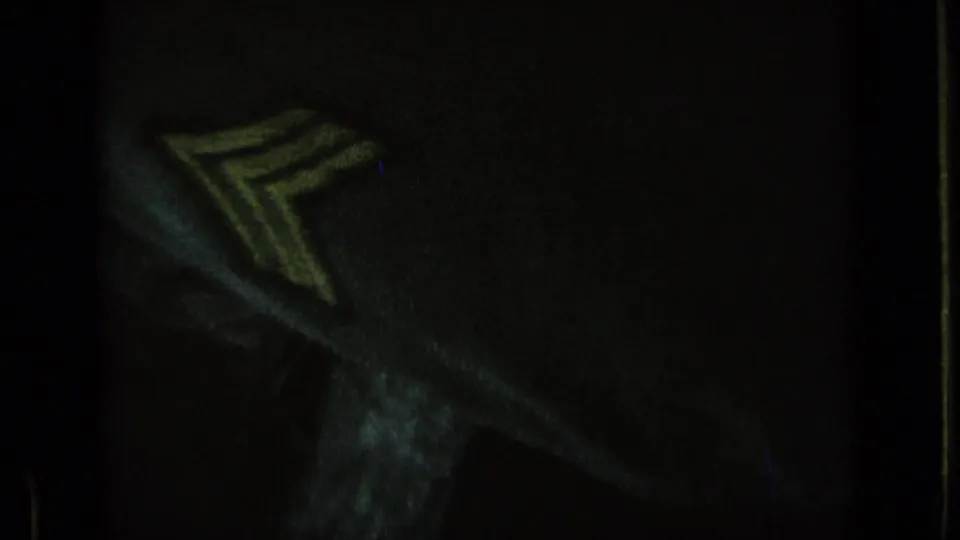 Underexposed 8mm frame showing part of a uniform sleeve with a chevron-style insignia, likely near the military vehicles.