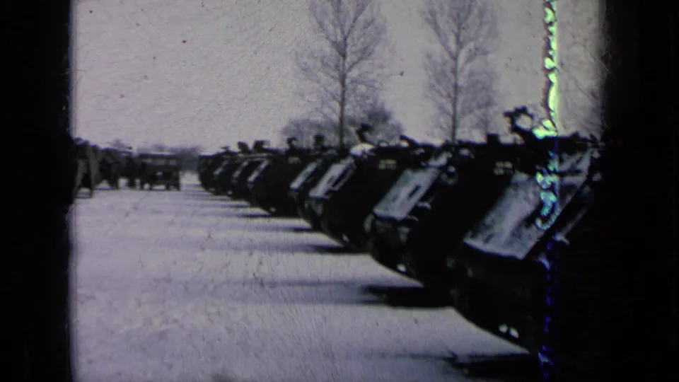 Parked tanks line a snowy field or base road, with a distant vehicle and bare winter trees visible beyond the row.