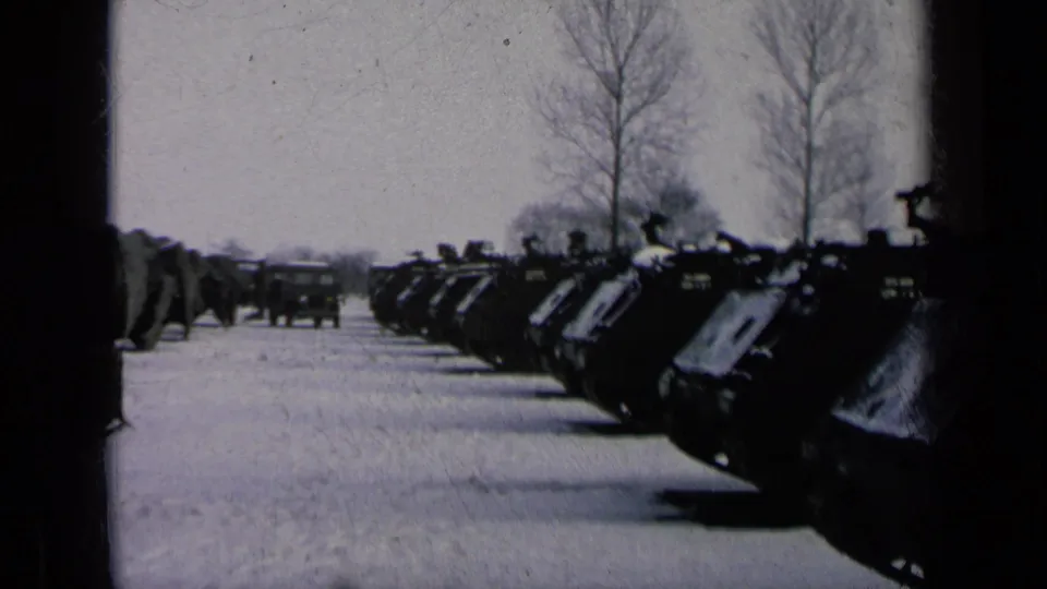 Armored vehicles stand in formation along a snow-covered base road, with bare trees and a truck visible ahead.