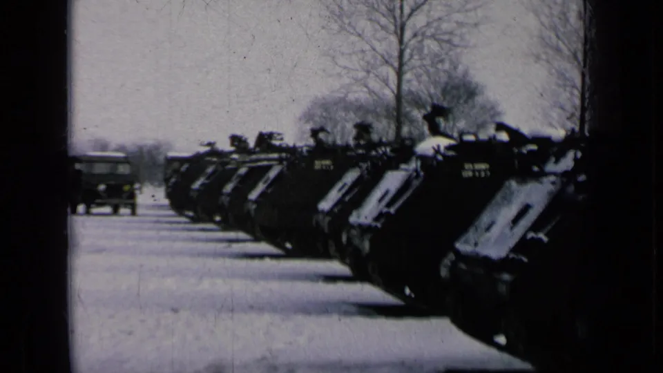 A military truck moves along a snow-covered road beside rows of armored vehicles at a winter base.