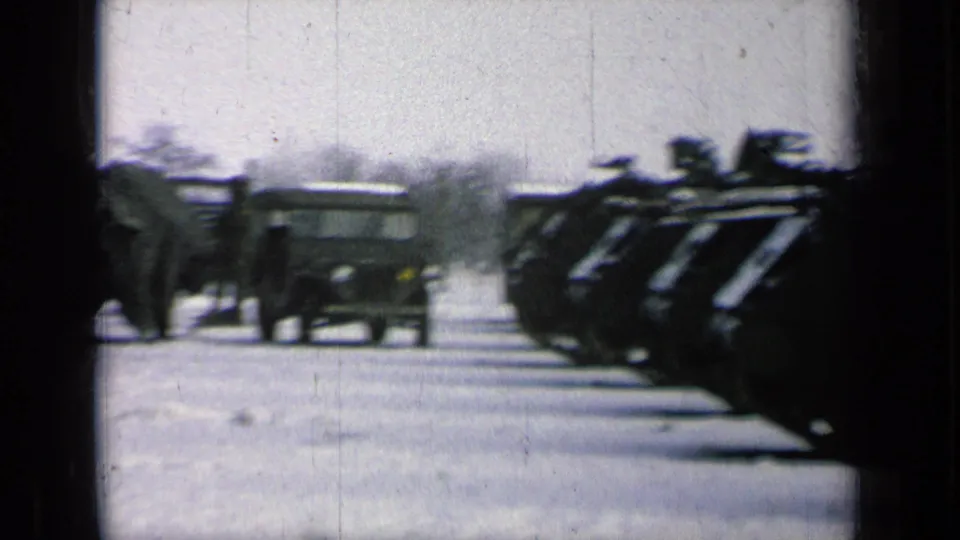 Wide 8mm frame of U.S. Army armored vehicles lined along a paved area, with a small utility vehicle and bare trees nearby.