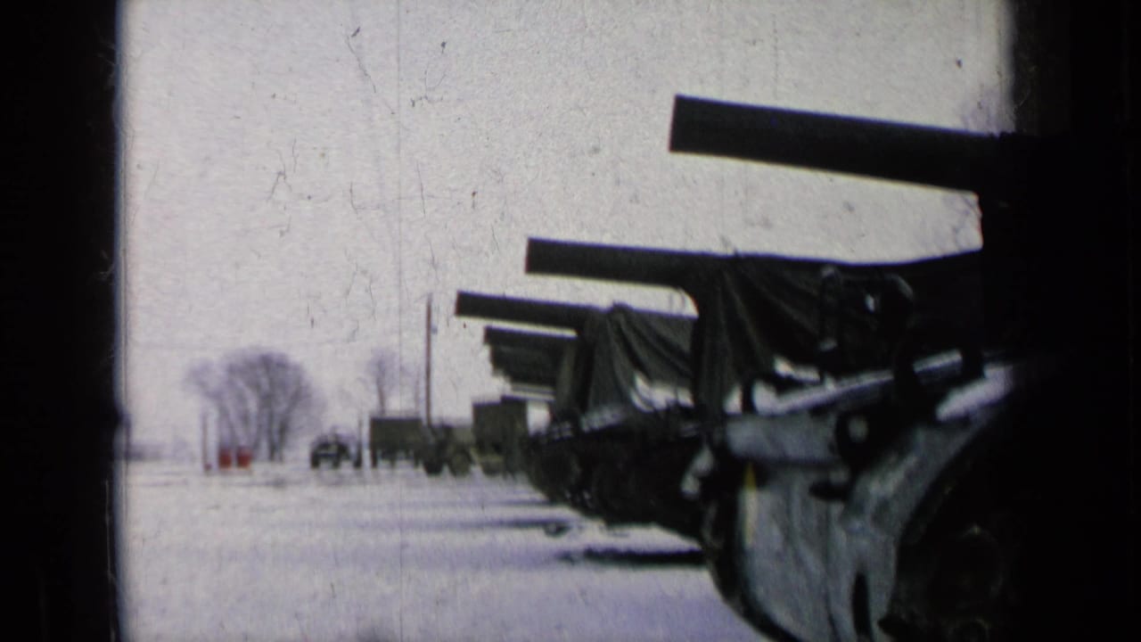 1963 8mm frame of covered artillery lined beside snow, with military trucks and bare trees in the distance.