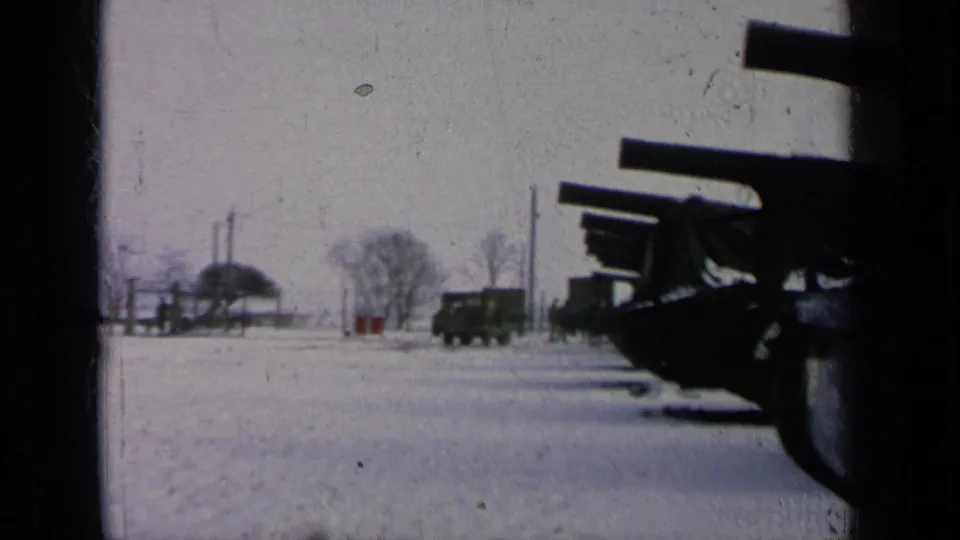 Snowy base view with a covered artillery row on the right and dark military trucks near bare winter trees.