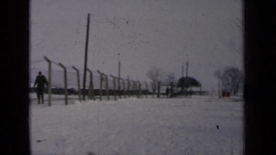 A distant figure walks near fence posts across a snow-covered military base area under gray winter light.