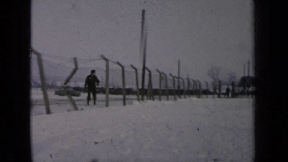 A uniformed man walks across a snowy military-base perimeter in Germany, 1963, with barbed wire and distant buildings behind him.