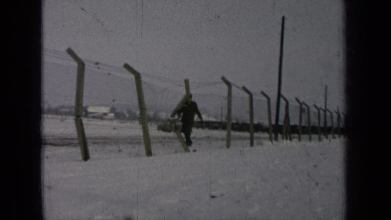 Man in dark uniform walks near a barbed-wire fence in snow.