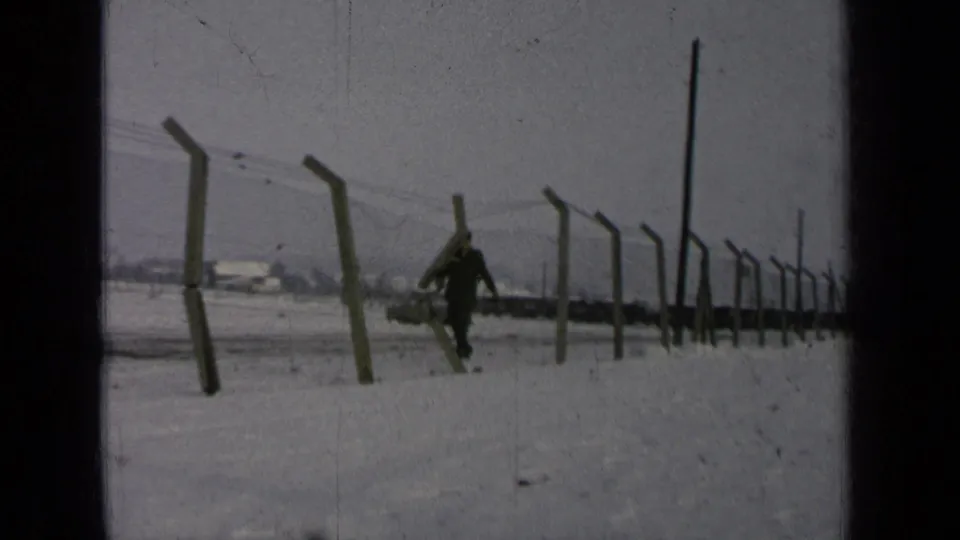 The uniformed man moves closer along the snowy fence line at a 1963 Germany military base, with low buildings in the distance.