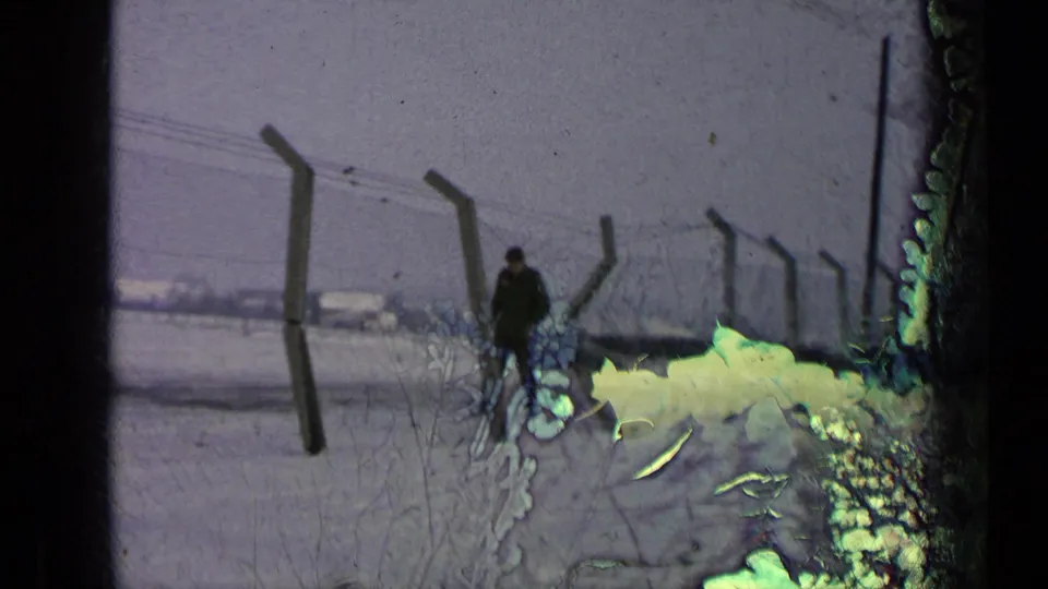 A dark-uniformed man is seen beside angled fence posts and barbed wire on snow-covered ground, with 8mm film damage visible across the frame.