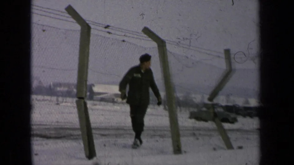 The man in uniform passes close to the camera near barbed-wire fencing, with snow, a parked car, and distant buildings in view.