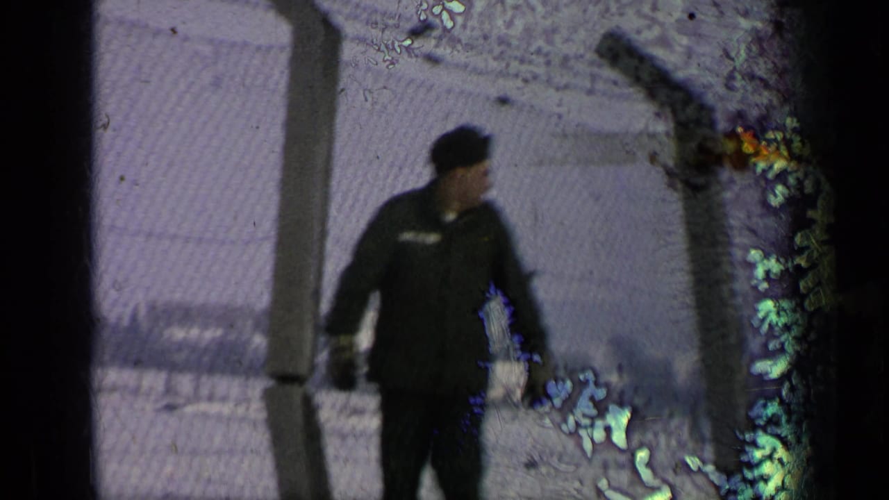 A uniformed man in a dark cap walks past a chain-link fence in snowy conditions, from 1963 Germany military-base footage per metadata.
