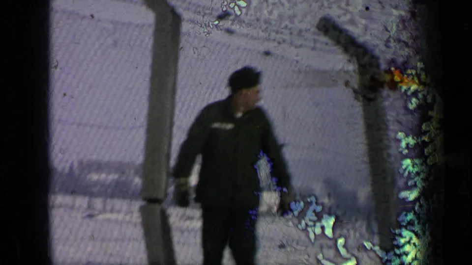 A uniformed man in a dark cap walks past a chain-link fence in snowy conditions, from 1963 Germany military-base footage per metadata.