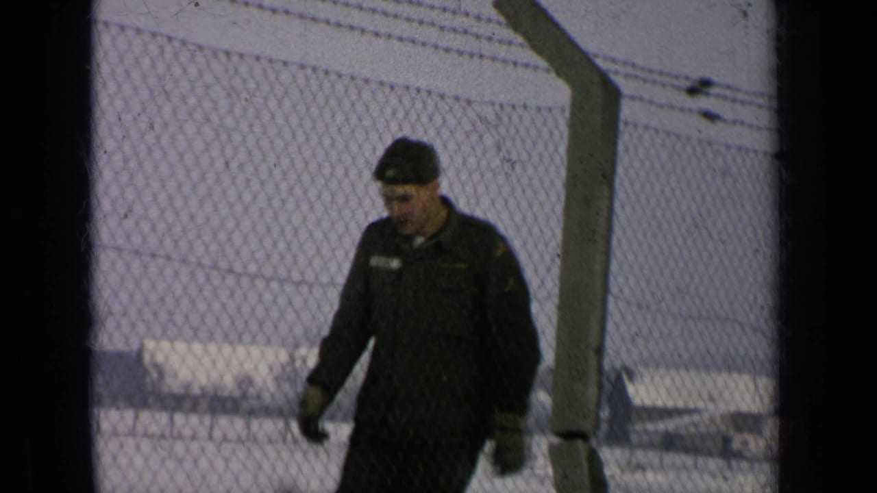 Uniformed man walks near a fence with snow and low buildings behind him.