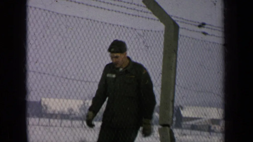 The soldier continues along the chain-link fence, with snow on the ground and pale buildings visible beyond the perimeter.