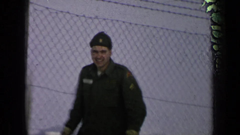The soldier appears closer to camera and smiles faintly while walking past the snowy chain-link fence.