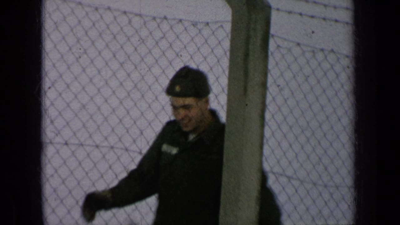 Uniformed soldier smiling and waving behind a chain-link fence.