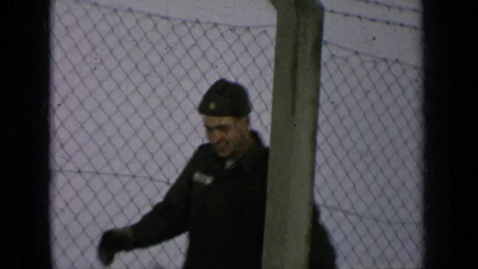 A U.S. Army soldier in dark uniform and cap smiles toward the camera behind a chain-link fence in metadata-supported 1963 Germany footage.