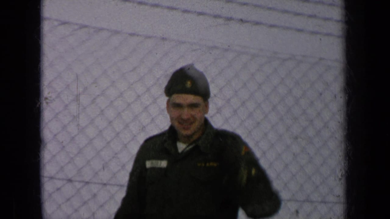 Uniformed soldier facing the camera beside a chain-link fence.