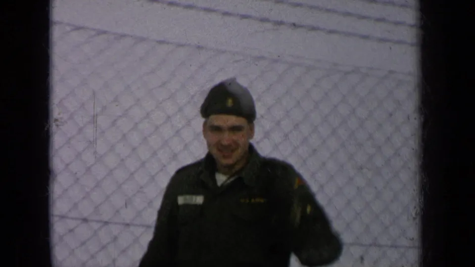 The soldier stands closer to center frame, facing the camera in dark military clothing with fence lines visible behind him.