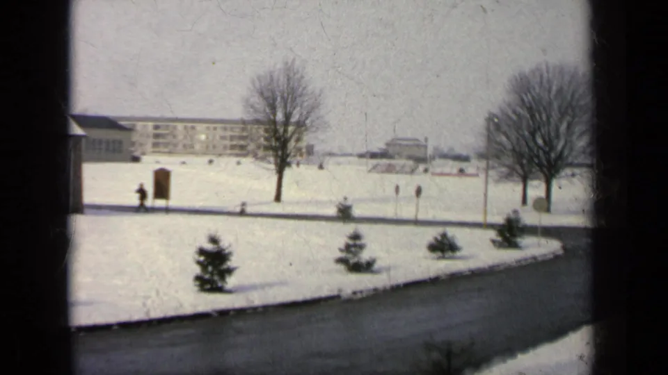 Snow-covered grounds on a reported Germany military base, seen from indoors, with a curved road, bare trees, evergreens, distant buildings, and a walker.