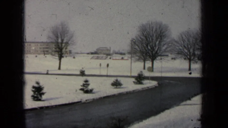 Winter exterior on a reported Germany military base, with snow-covered grounds, a curved road, small evergreens, bare trees, signs, and distant buildings.