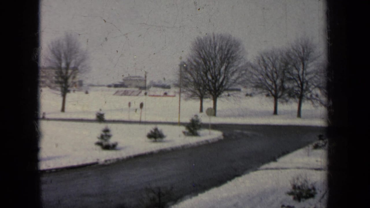 Snowy curved road seen through dark window framing with bare trees and distant low buildings.