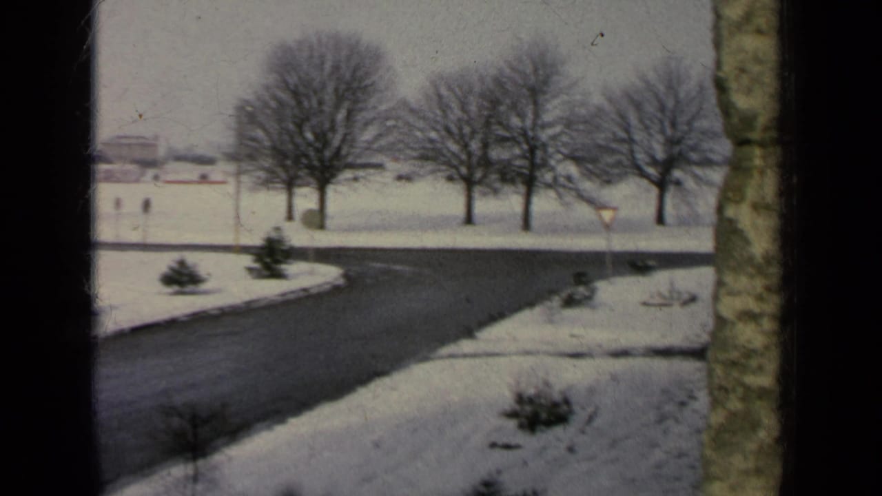 Snow-covered road curves past lawns, shrubs, bare trees, and low buildings in the distance.