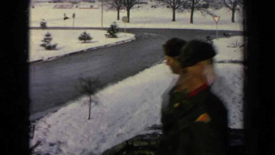 Two military-style uniformed figures pass near the window view of a snowy 1963 Germany base road.